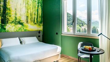Una camera d'albergo con un letto, un tavolino con la colazione, una sedia, una finestra vista foresta e montagne, e una parete verde.