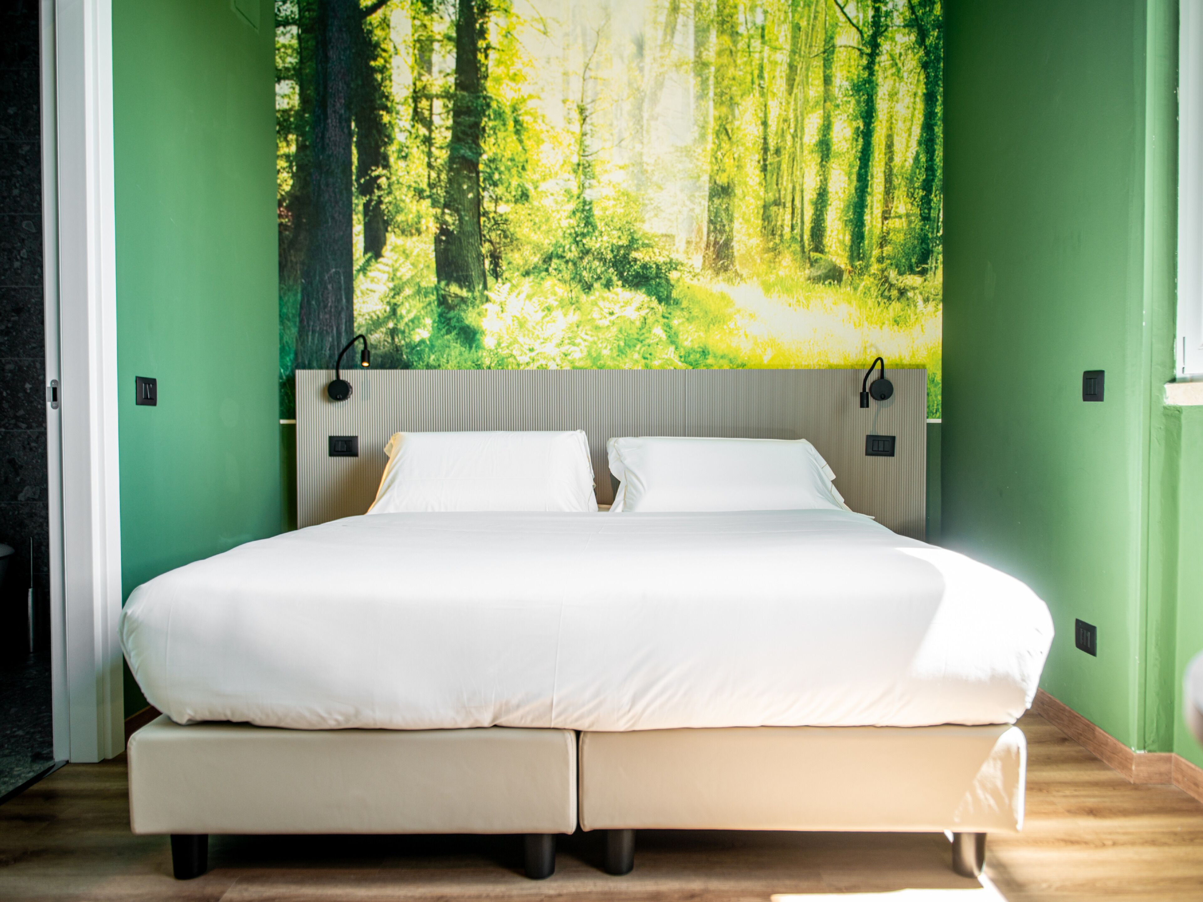 Ein Bett mit weißer Bettwäsche, ein Kopfteil mit Waldgemälde und grüne Wände.
