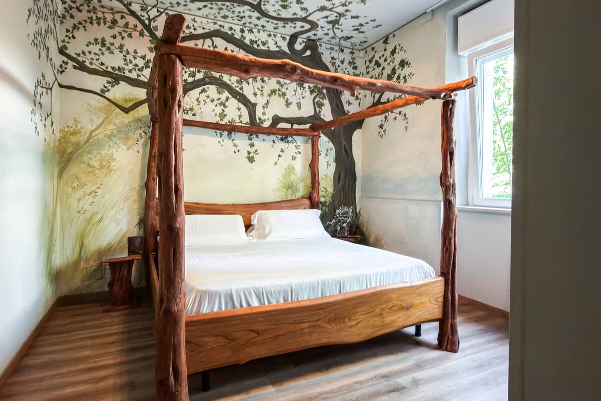 Ein hölzernes Himmelbett mit weißer Matratze und Kissen vor einer Baum-Mural.