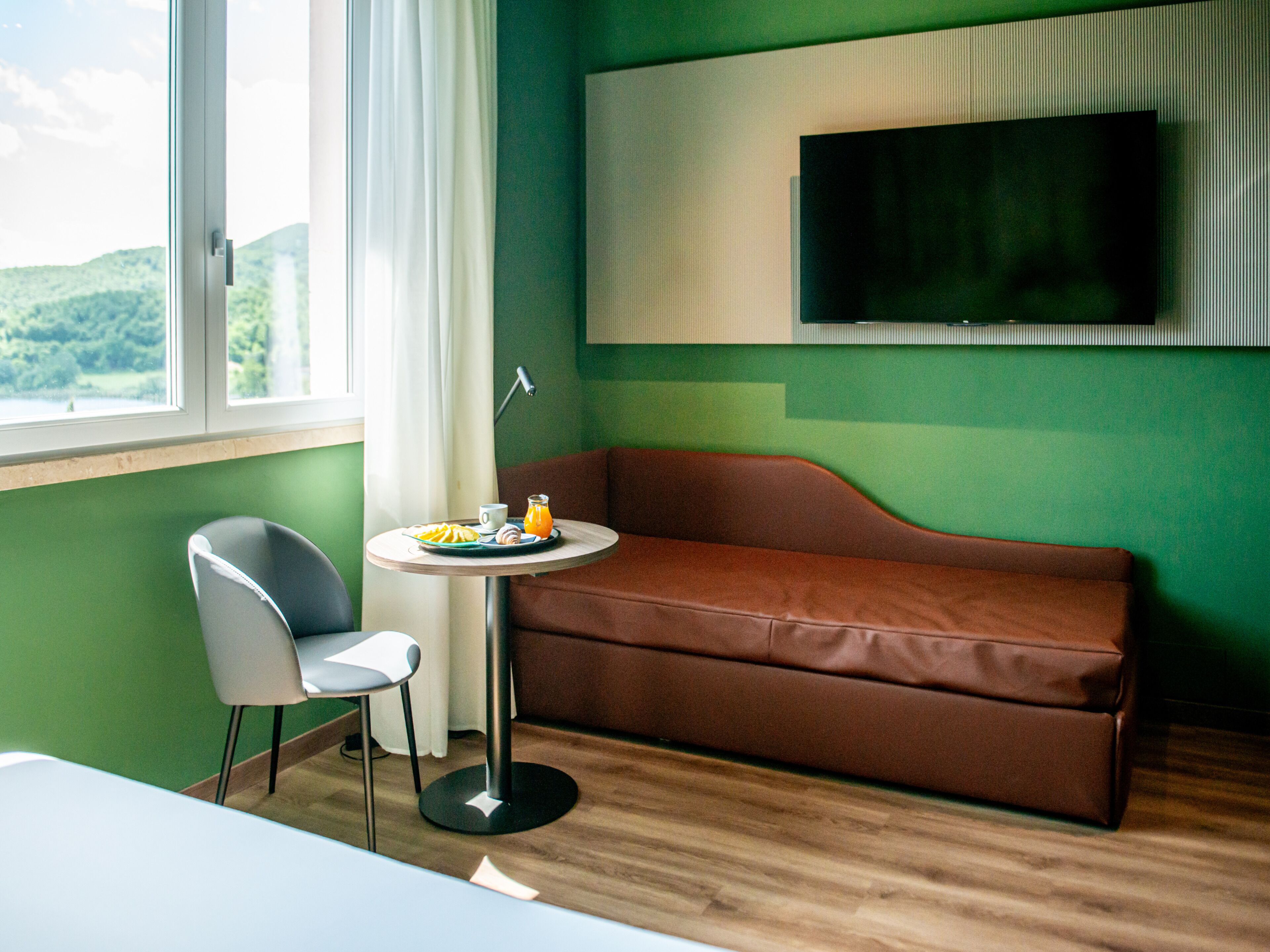 A room with a brown sofa, a small round table with a tray of food, a chair, a TV mounted on the wall, and a window with a view of greenery.