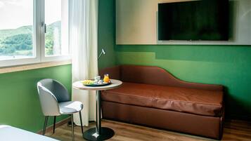 A room with a brown sofa, a small round table with a tray of food, a chair, a TV mounted on the wall, and a window with a view of greenery.