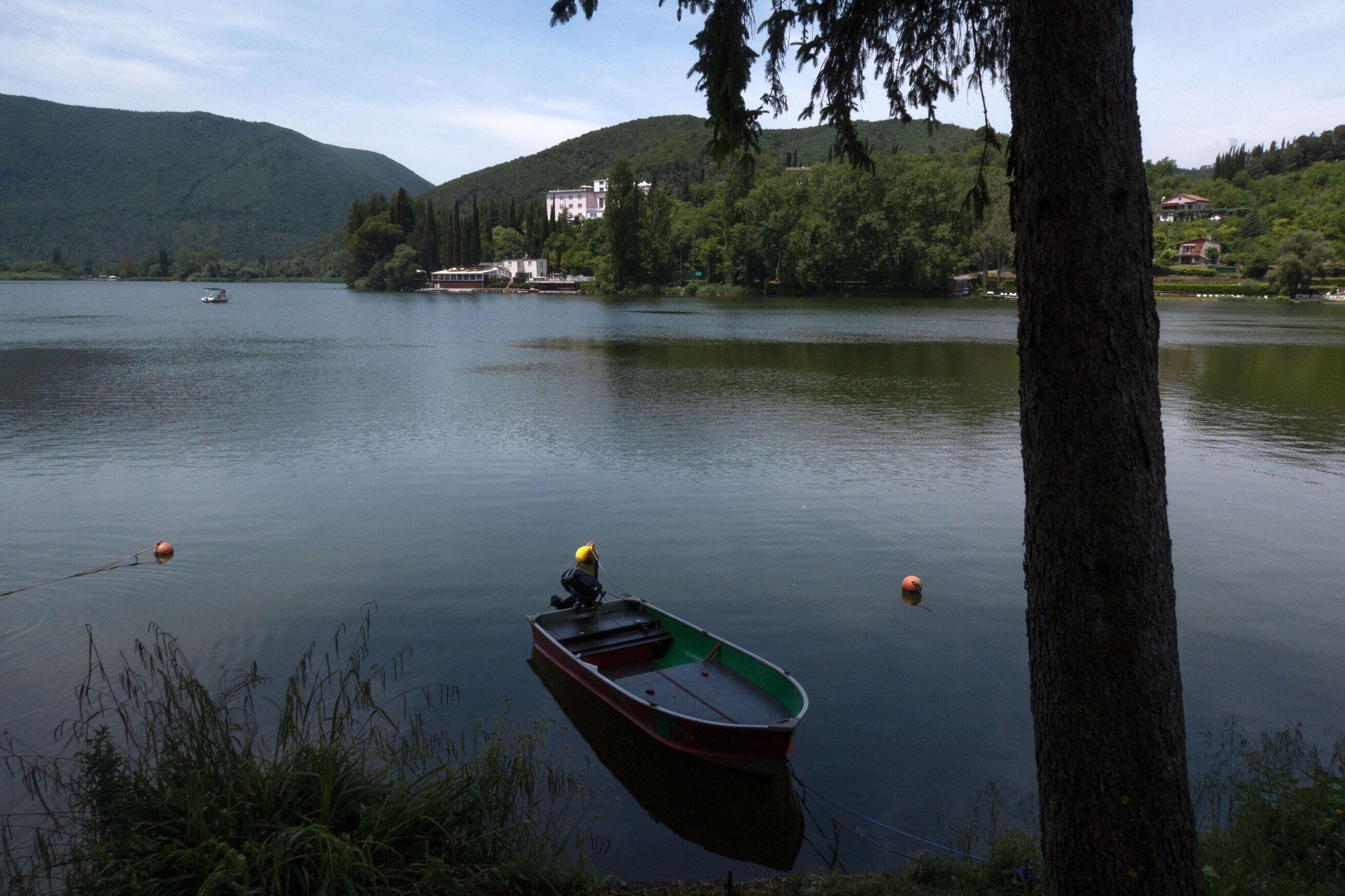 A calm lake with a small boat tied to a tree, surrounded by lush greenery and hills in the background.