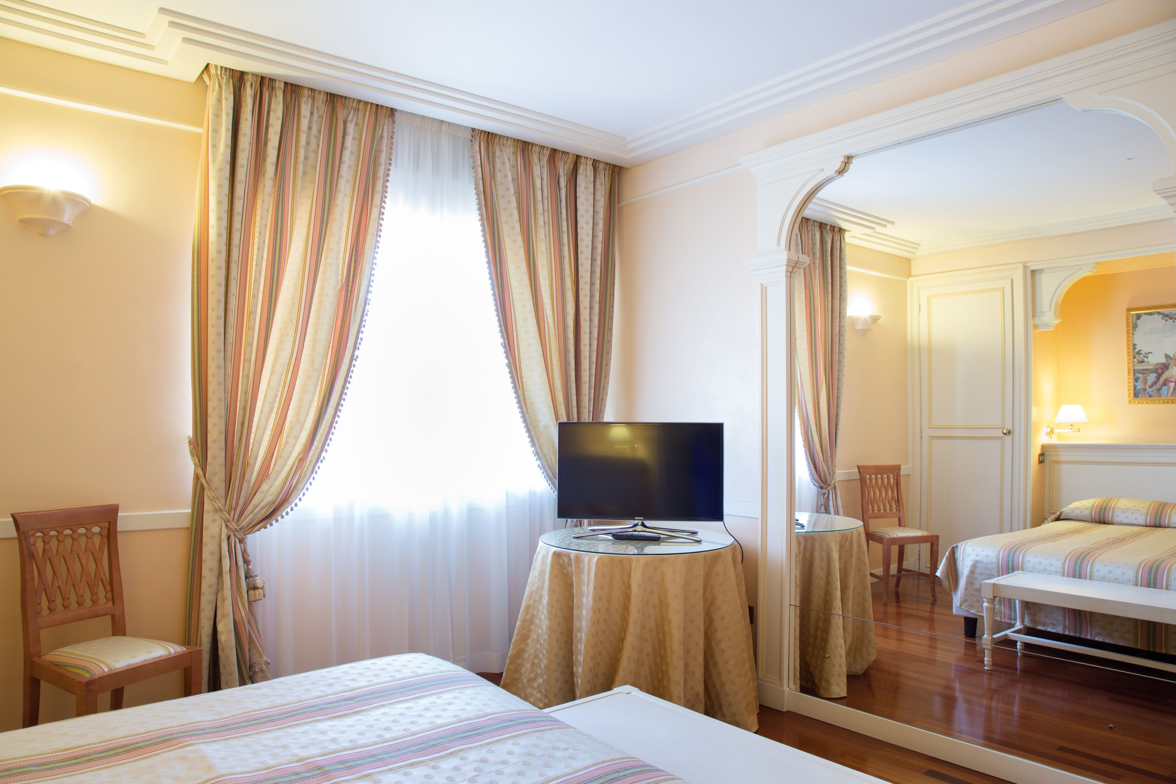 Una habitación de hotel con una cama, un televisor sobre una mesita, una silla y un espejo que refleja la habitación.