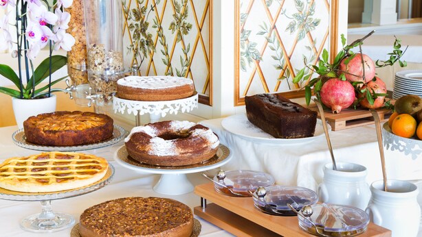 A variety of cakes and pastries on display, including a chocolate cake, a fruit tart, and a bundt cake, along with a vase of flowers and a fruit basket. - Tuder (Todi)