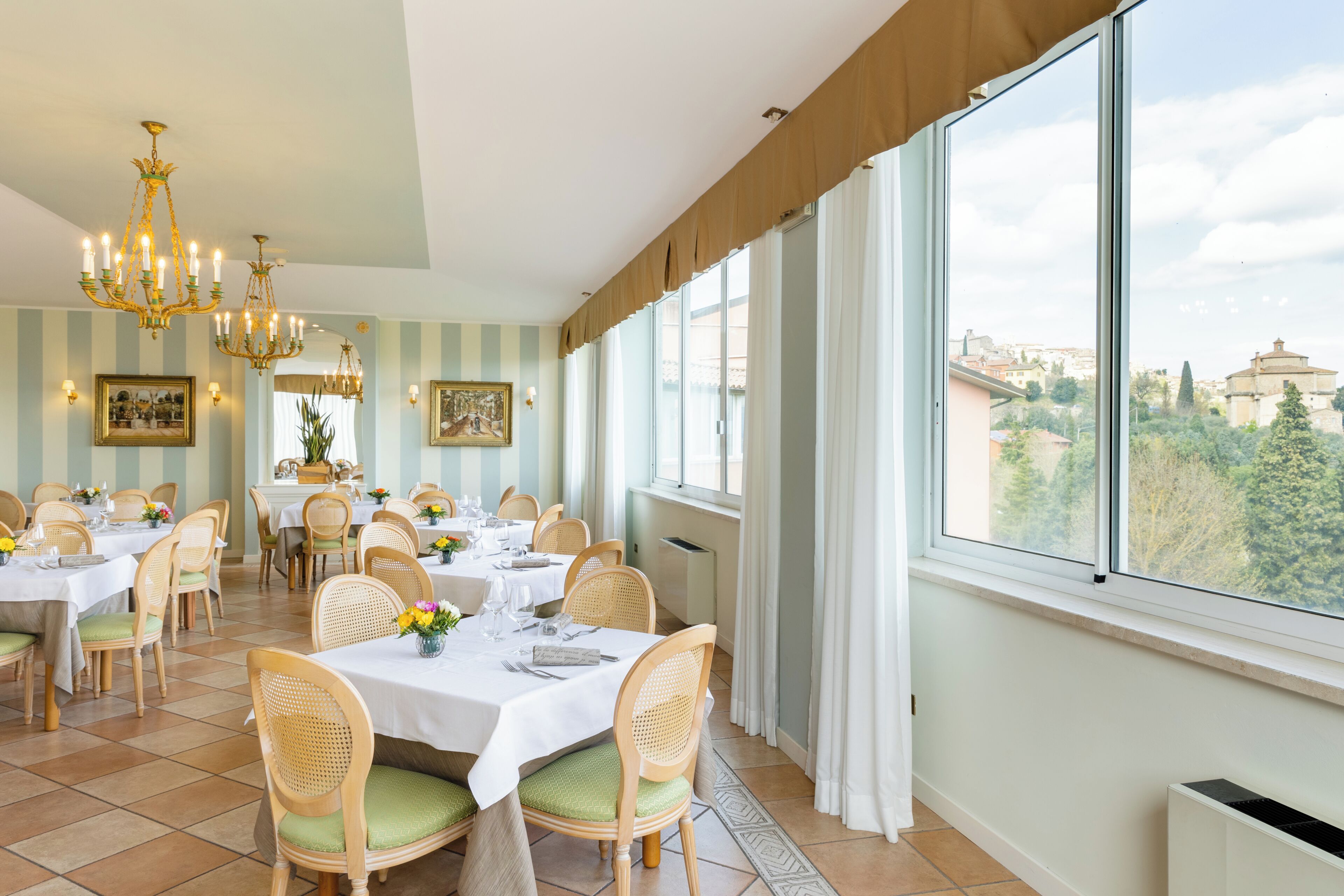 A dining area with tables set for a meal, large windows with a view of buildings, and a chandelier.