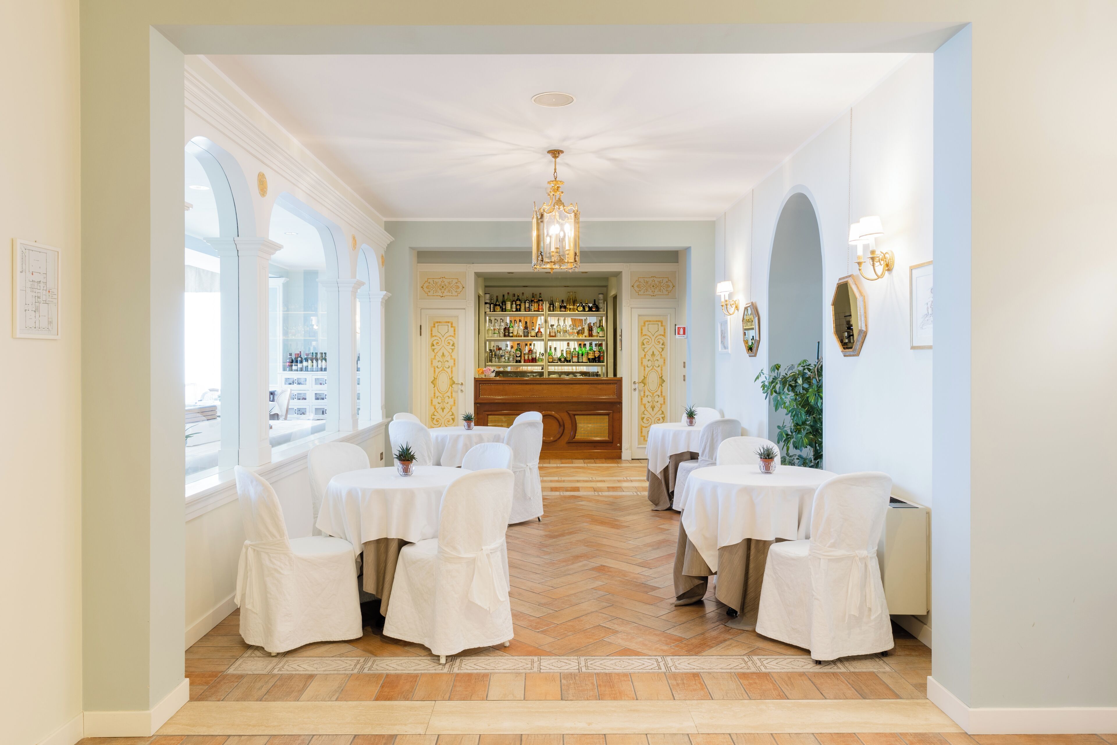 A restaurant with white chairs and tables, a chandelier, and a bar area with shelves of bottles.