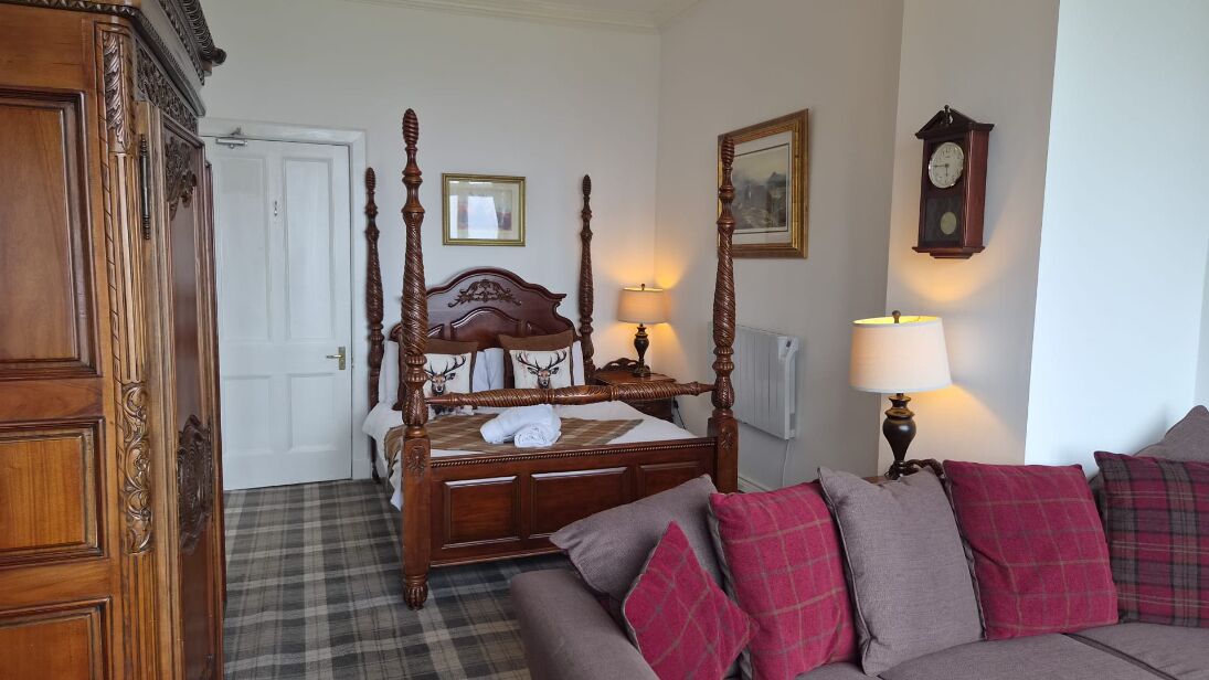 A bedroom with a four-poster bed, a wooden wardrobe, a plaid floor, a sofa with plaid cushions, a wall clock, and a framed picture.