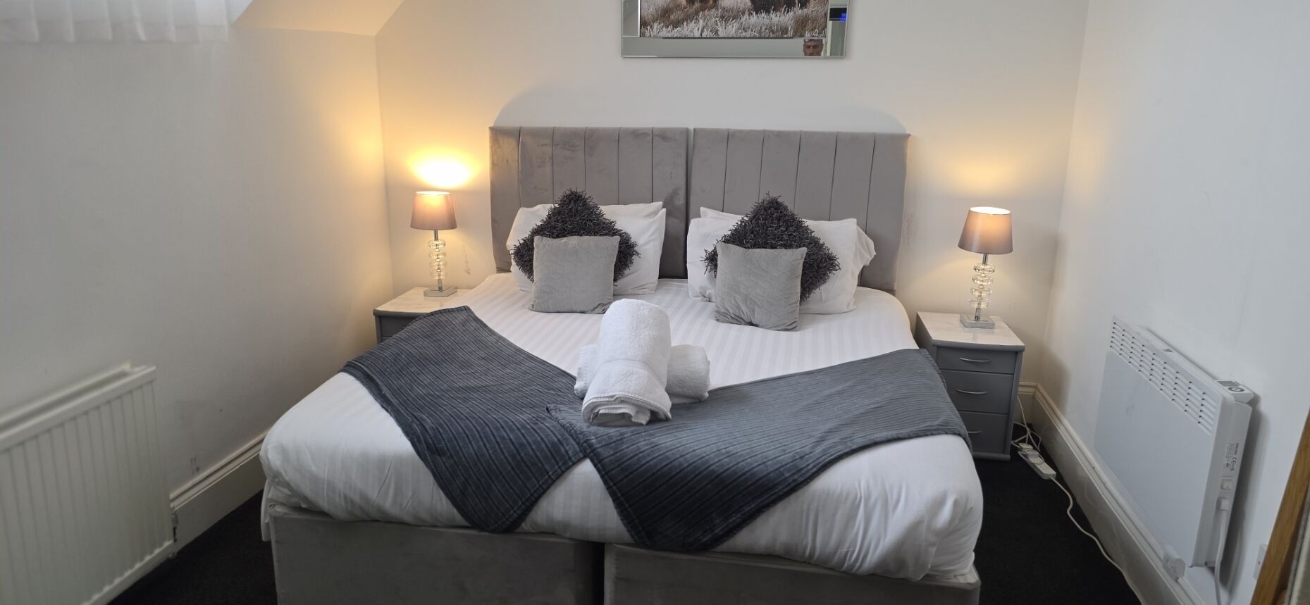 A neatly made bed with a grey headboard, two bedside lamps, and a framed picture on the wall.