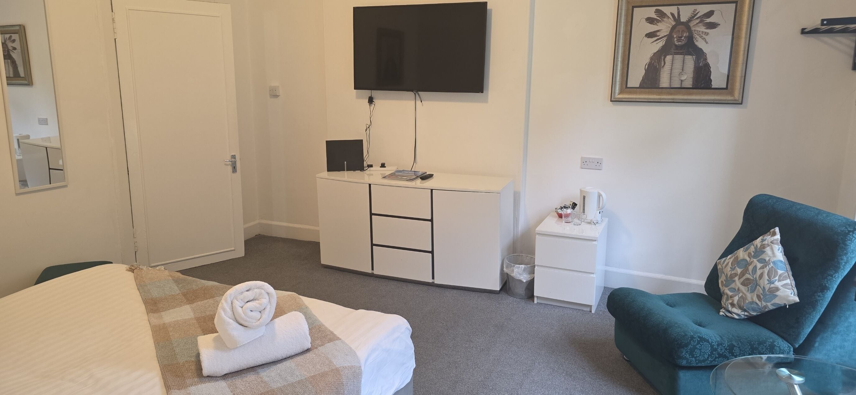 A hotel room with a bed, a desk with a flat-screen TV, a small white cabinet, a green armchair, and a framed picture on the wall.