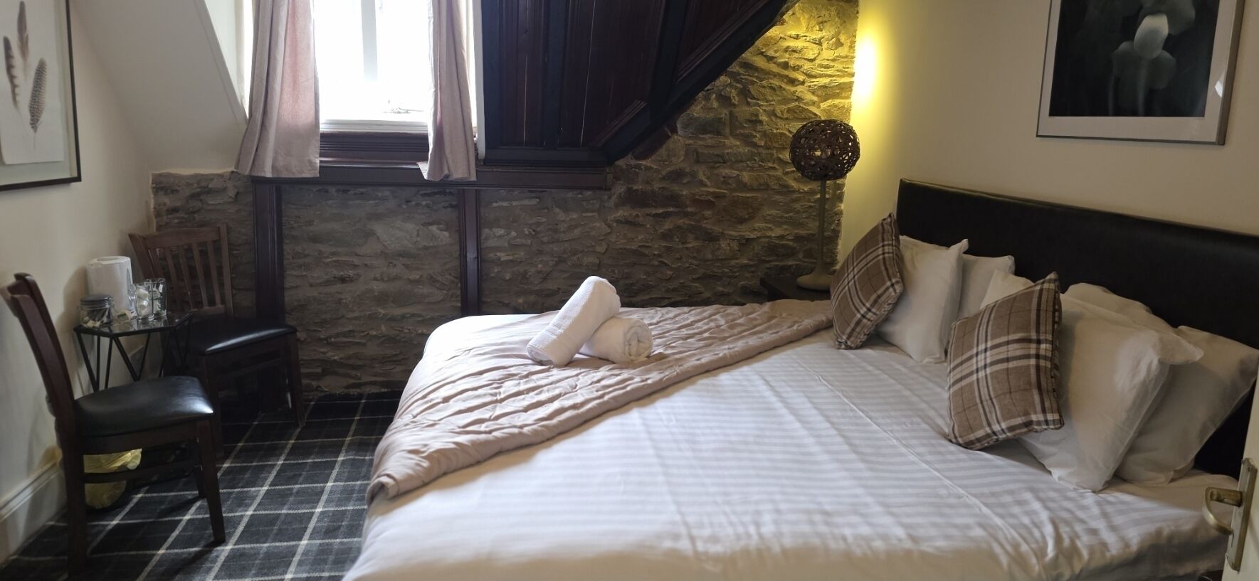 Un dormitorio con cama, mesita de noche, silla, lámpara y pared de piedra.