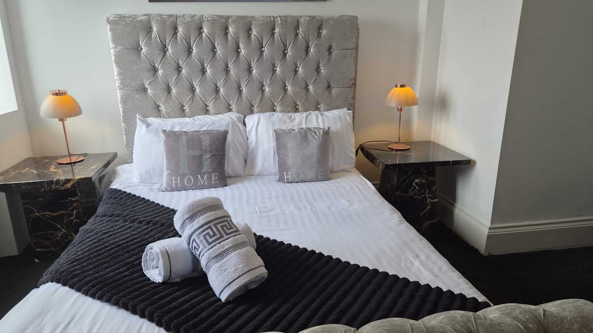Een net opgemaakt bed met een gestoffeerd hoofdbord, twee nachtkastjes met lampen en decoratieve kussens met de tekst 'HOME'.