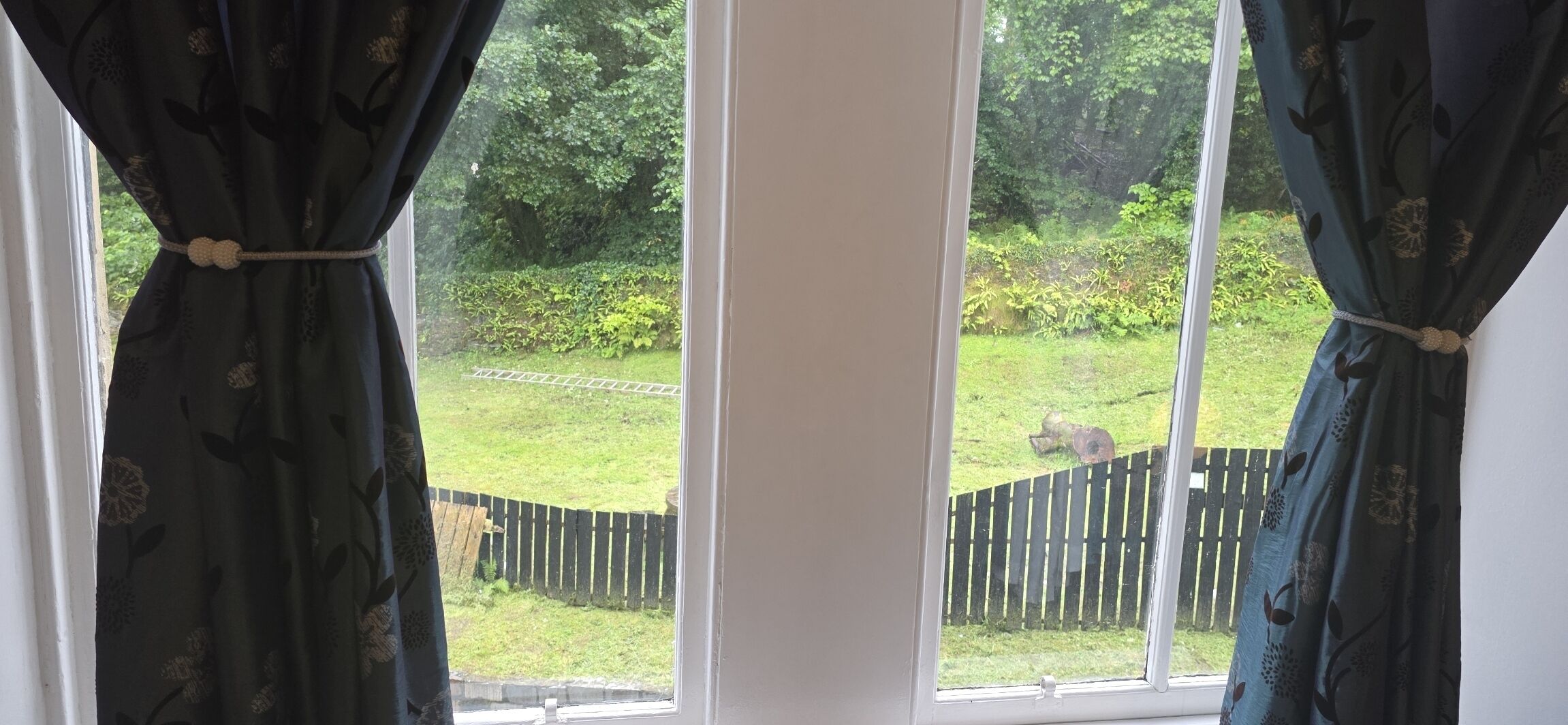 Una ventana con cortinas y vistas a un jardín, con una cerca de madera.