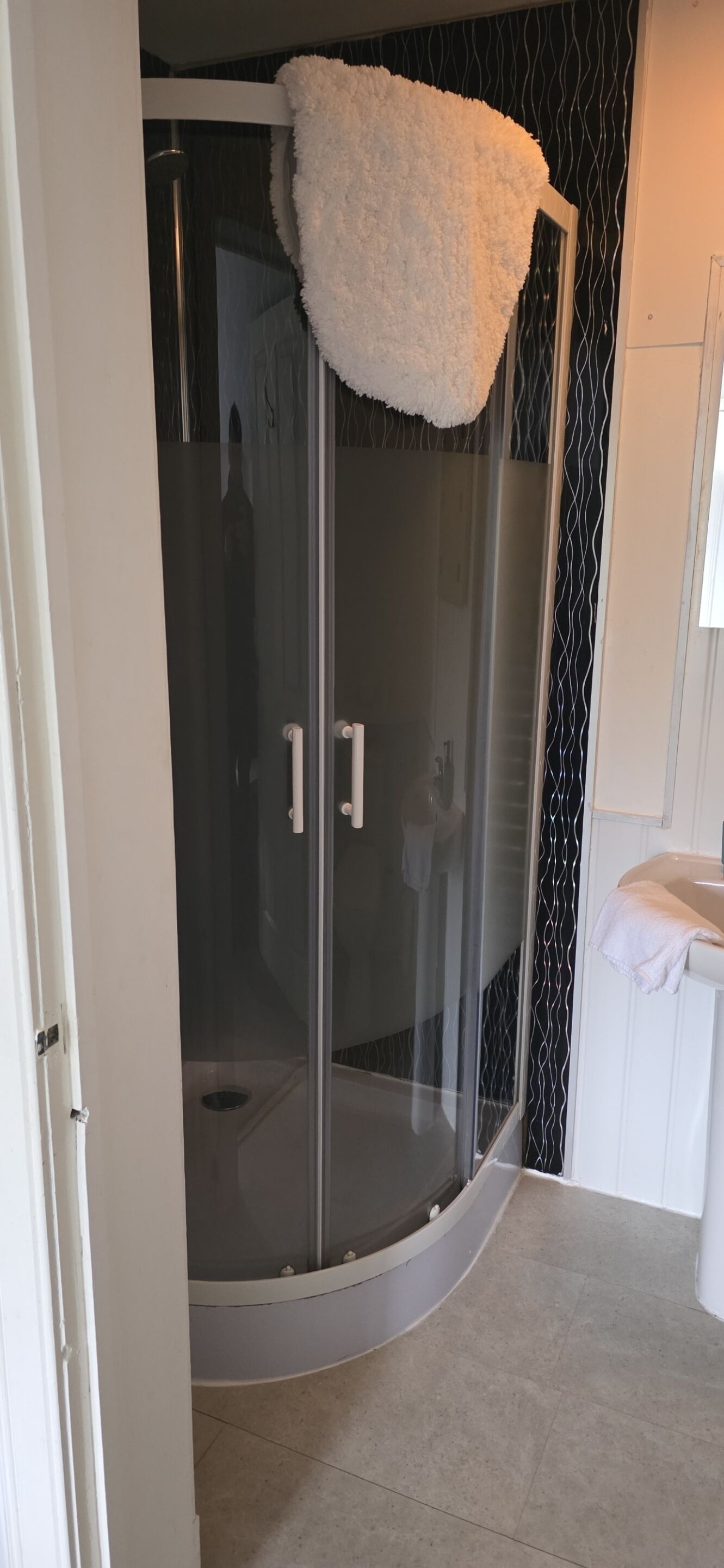 A glass-enclosed shower with a white fluffy bath mat, a white sink, and a patterned wall.