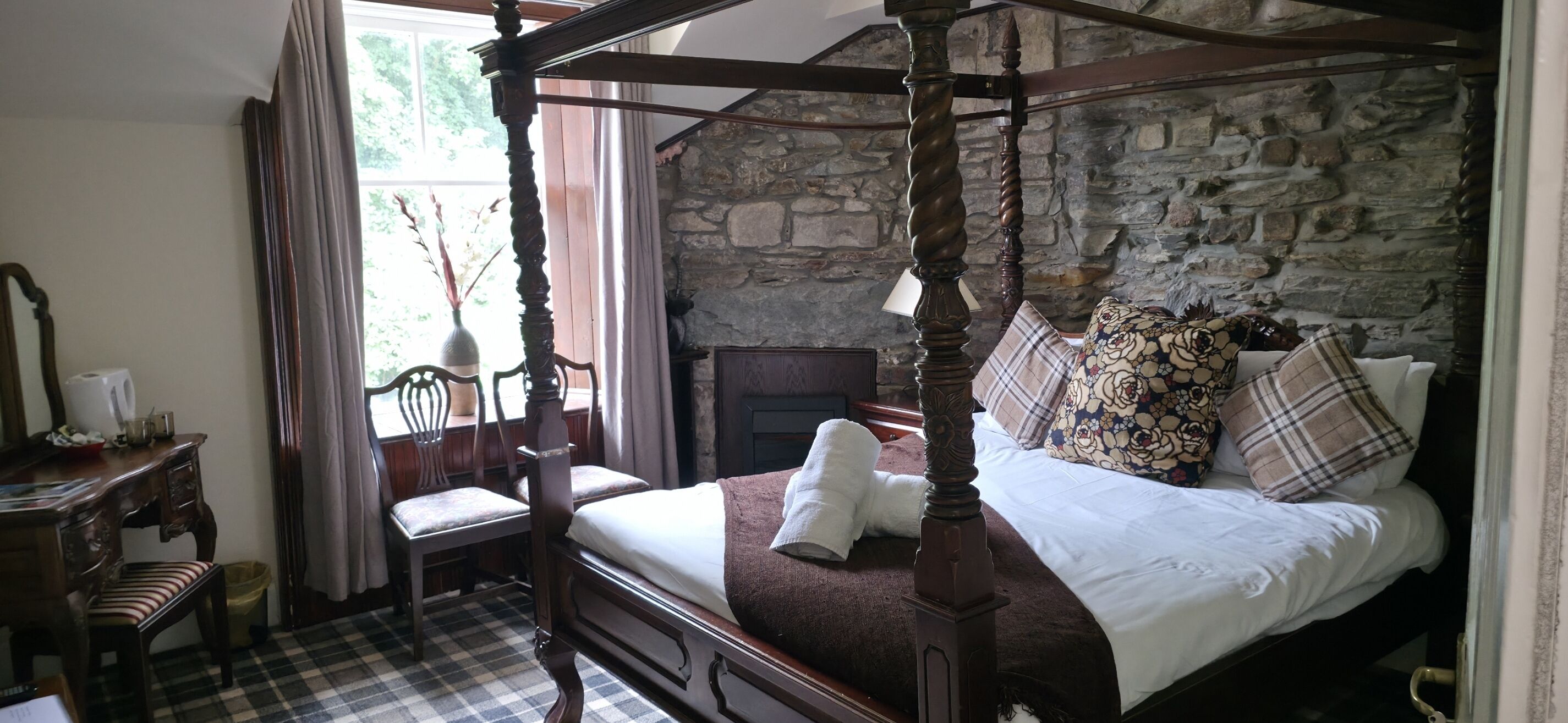 A four-poster bed with a stone wall backdrop, a wooden chair, a checkered floor, and a window with curtains.