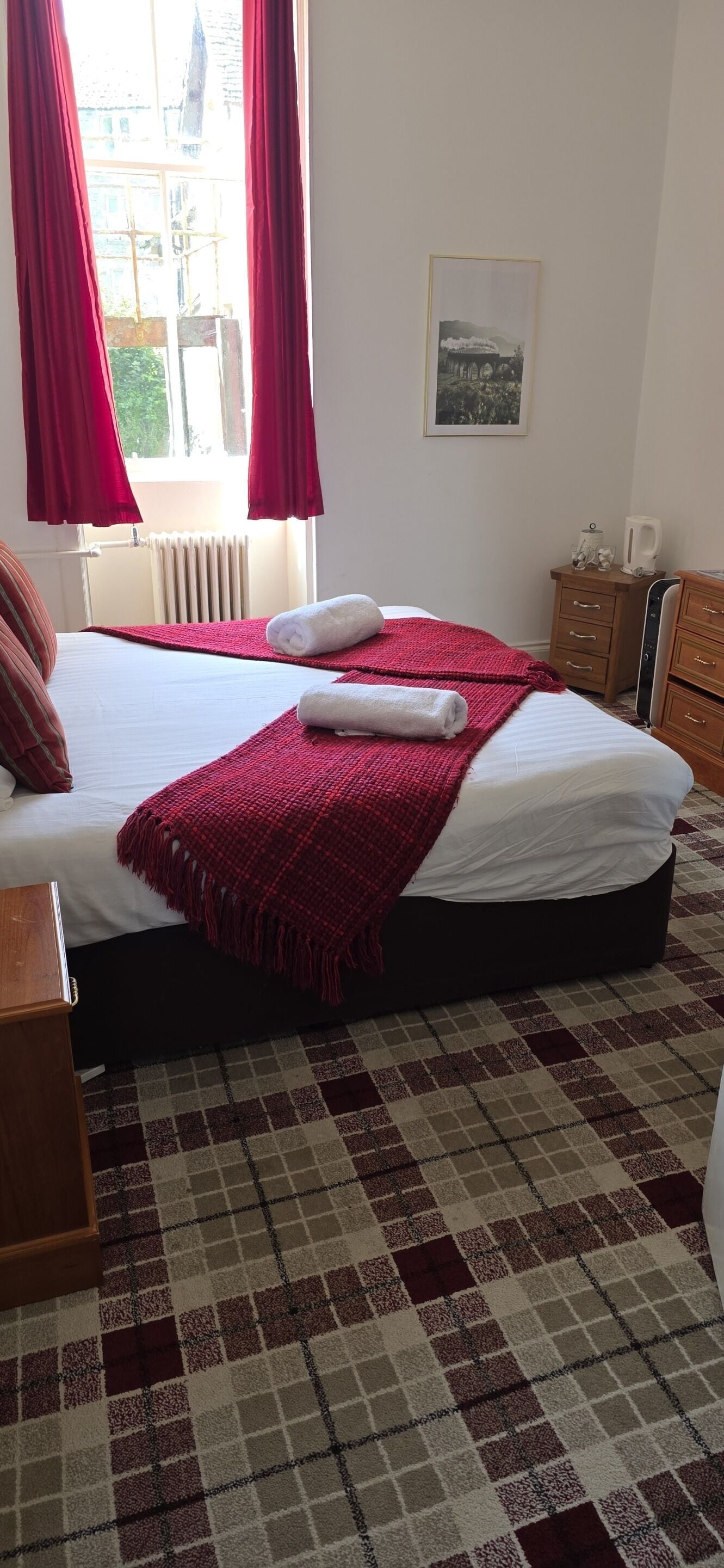 Quarto de hotel com cama, radiador, janela com cortinas, um quadro na parede e carpete estampado.