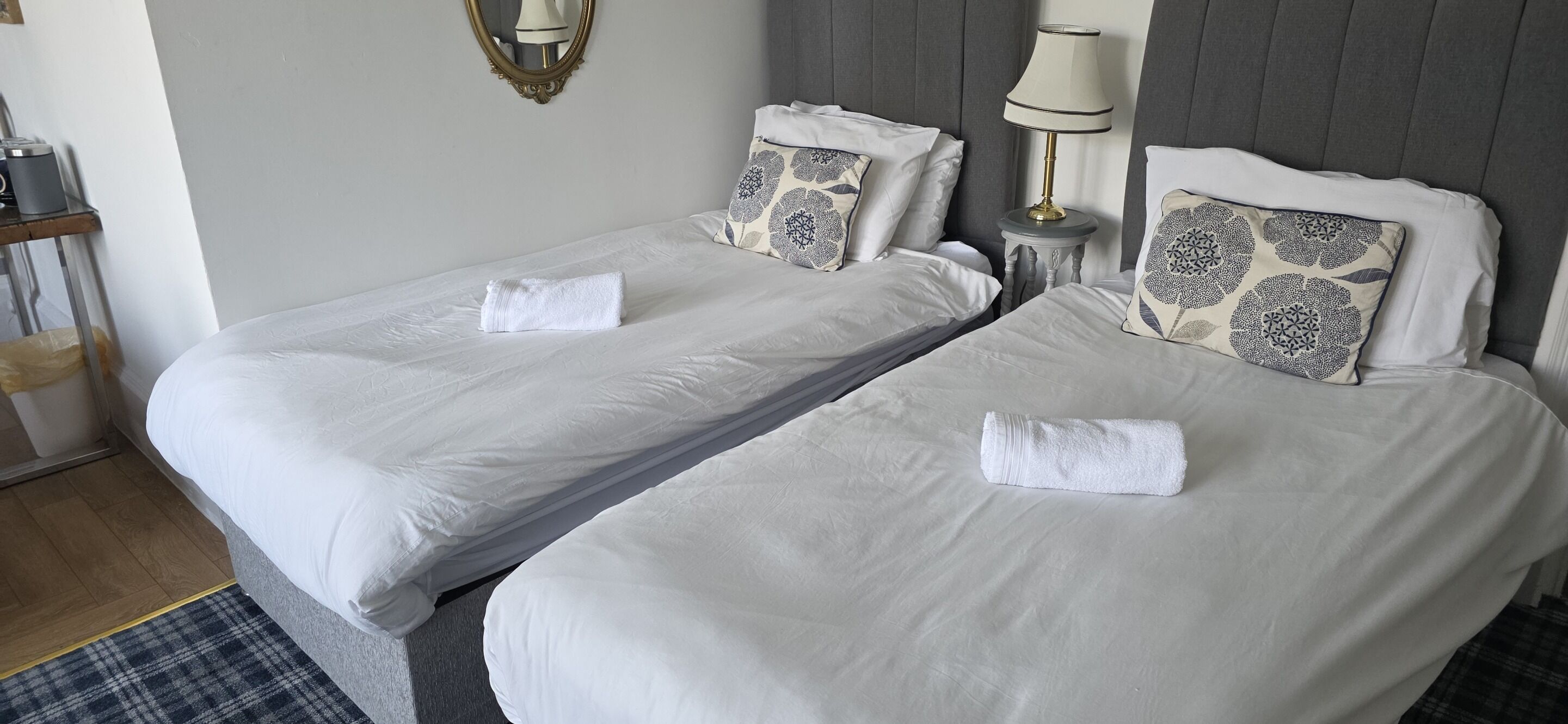 Deux lits simples avec du linge de lit blanc, une table de chevet avec une lampe et un miroir au mur.