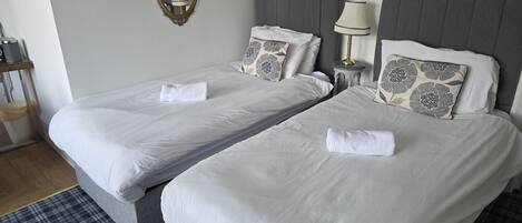 Deux lits simples avec du linge de lit blanc, une table de chevet avec une lampe et un miroir au mur.