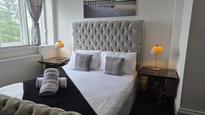 A neatly made bed with a tufted headboard, two bedside lamps, and a view of a pier through the window. - Royal Marine Hotel (Dunoon)