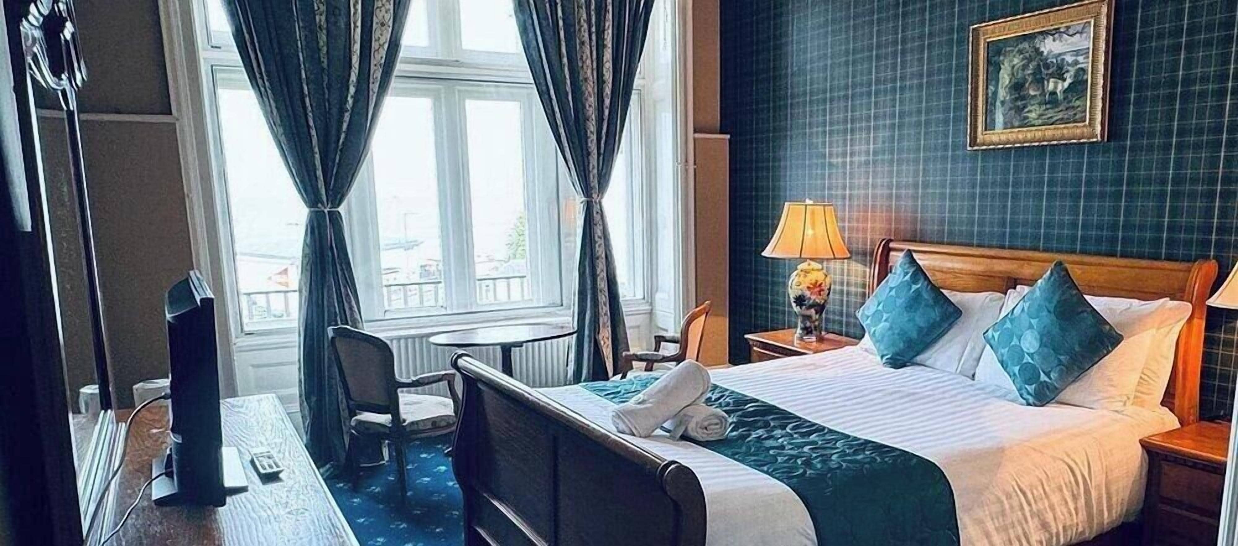 A hotel room with a bed, bedside table, lamp, and a view of the city through the window.