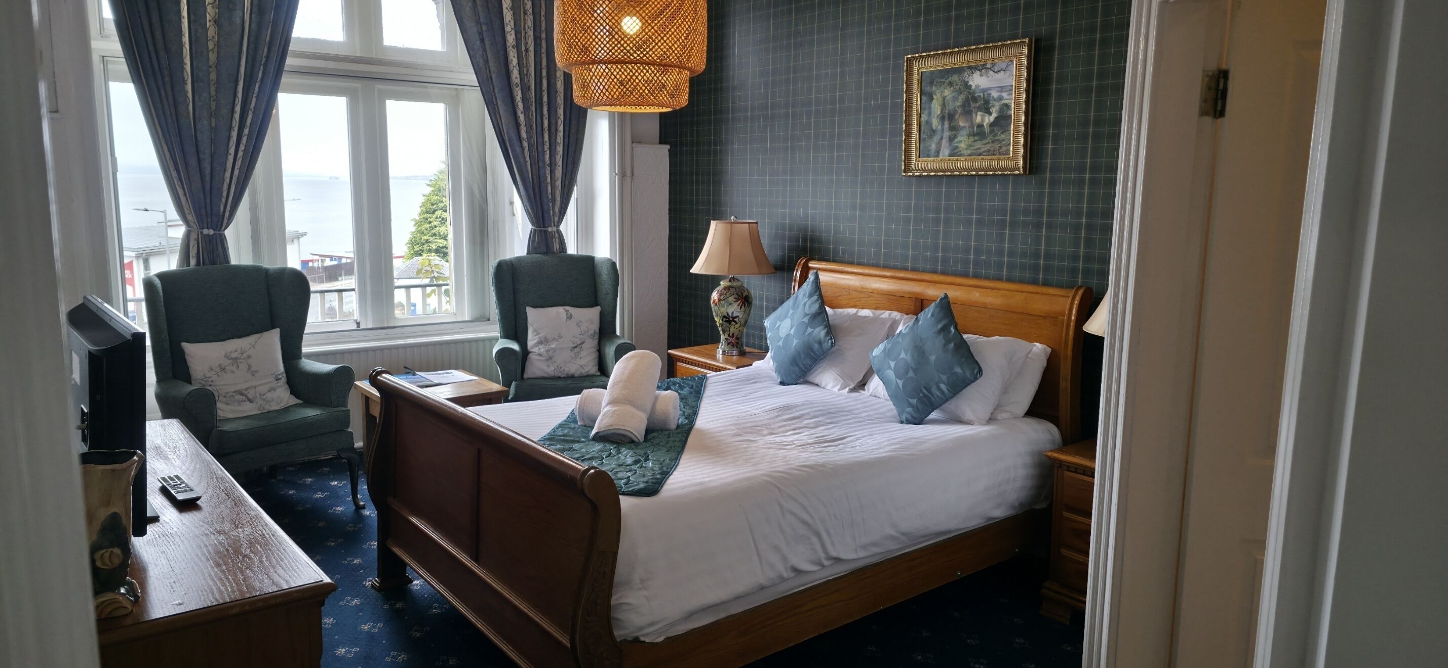 A hotel room with a bed, two chairs, a desk, a lamp, and a painting on the wall.