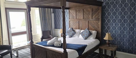 A four-poster bed with a wooden headboard, a bedside table with a lamp, and a chair in a hotel room with patterned wallpaper.