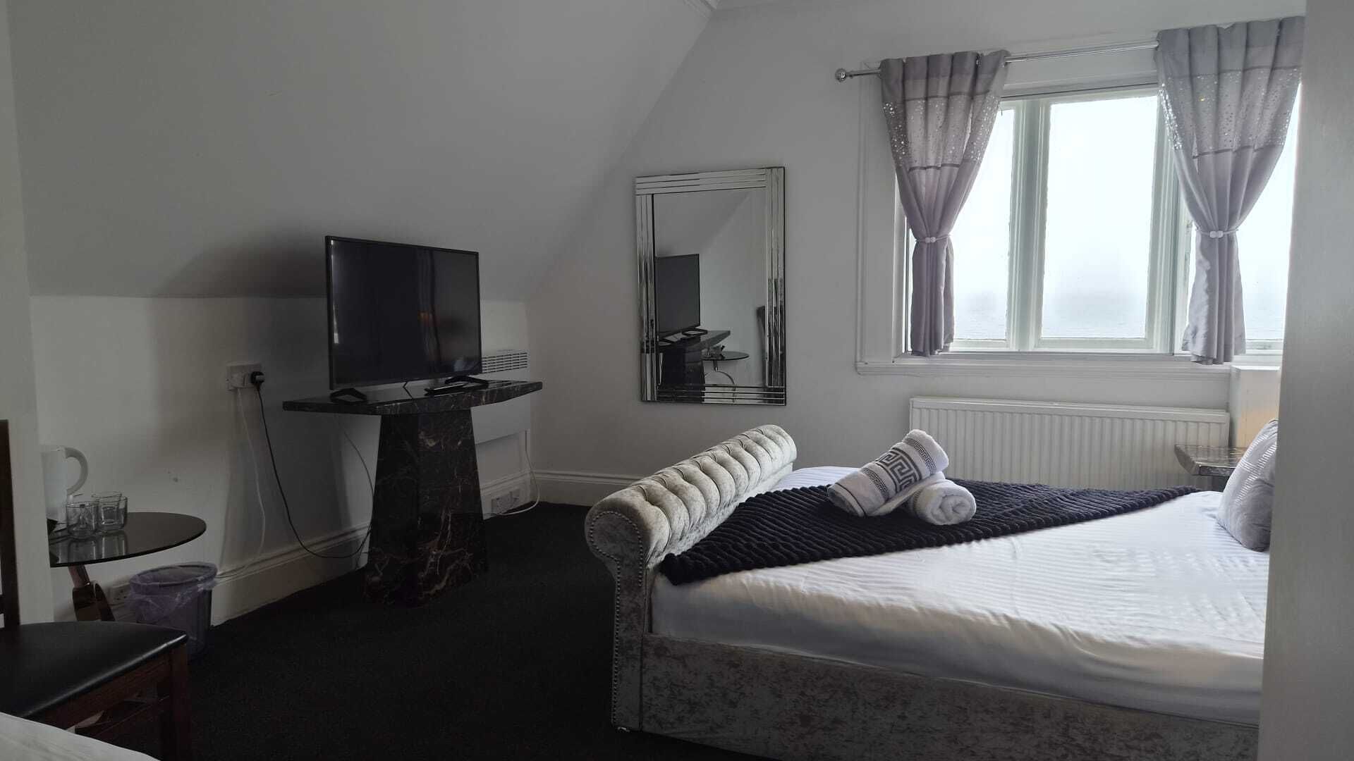 A hotel room with a bed, a television on a stand, a mirror, and a window with curtains.
