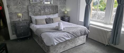 A neatly made bed with a white duvet, a fluffy pillow, and a folded towel on top. There are two bedside tables with lamps, a patterned wall, and a window with curtains.