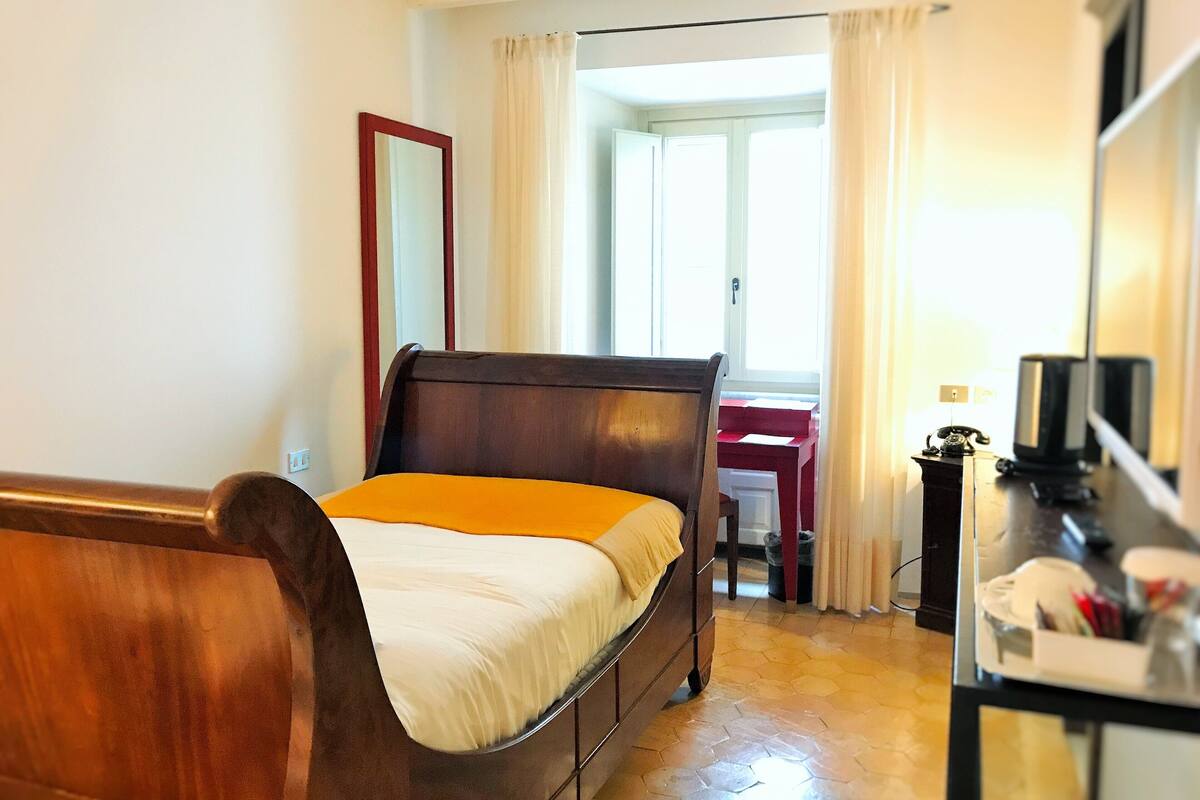 A bedroom with a wooden bed, a desk with a chair, a mirror, and a window with curtains.