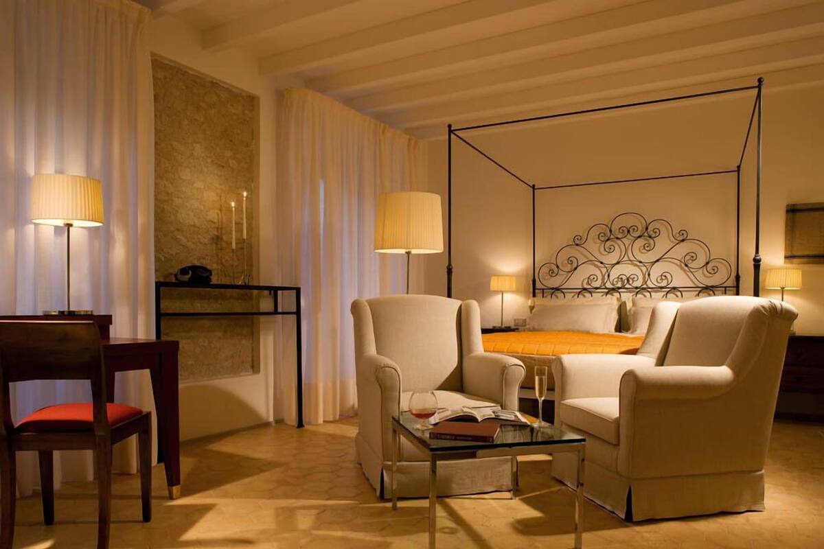 A hotel room with a four-poster bed, two armchairs, a small table, and a desk with a lamp.
