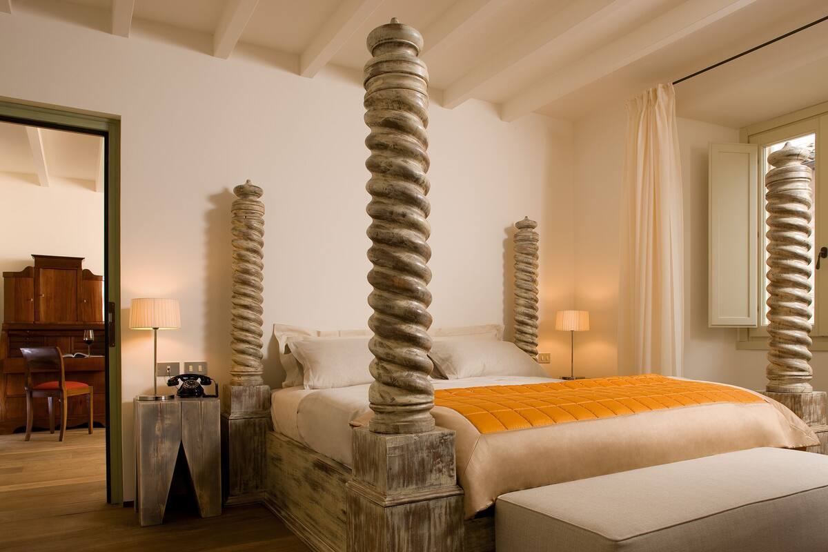 A four-poster bed with a wooden base, a bedside table with a lamp, and a chair in a room with wooden flooring and white walls.