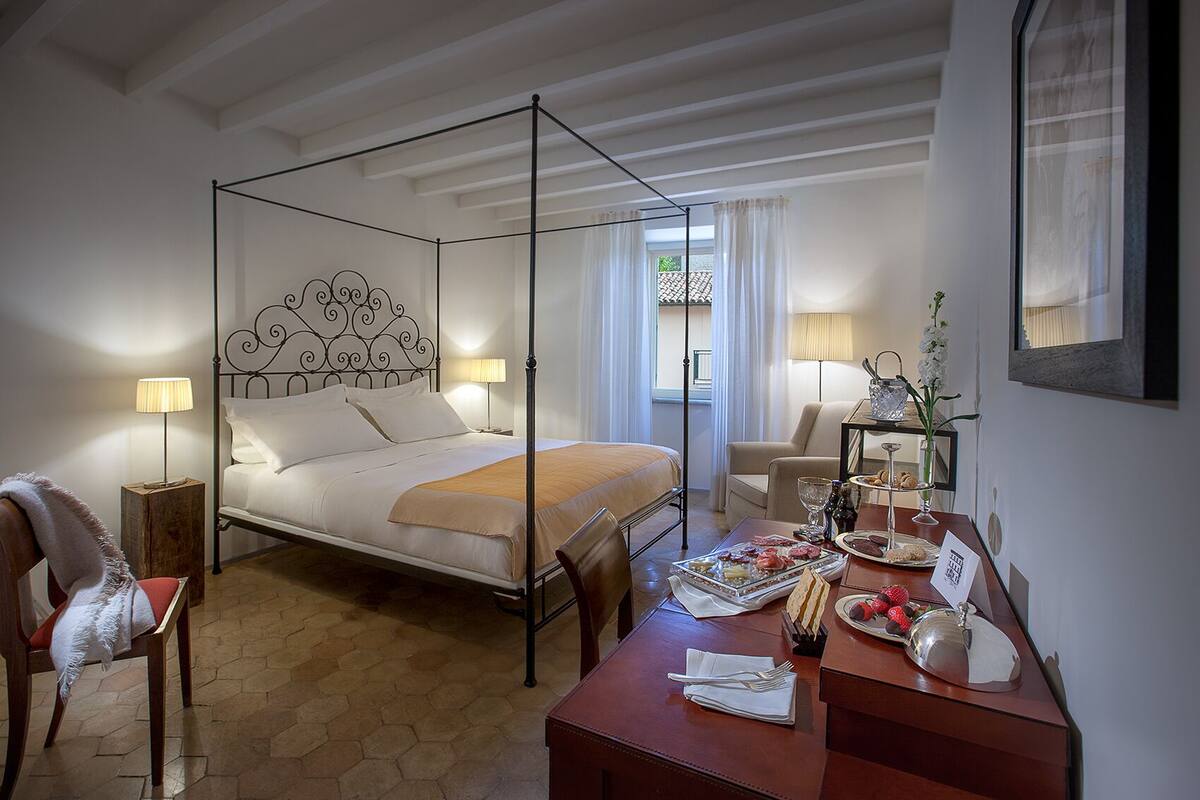 A four-poster bed with a decorative headboard, a wooden bedside table with a tray of food, a chair, and a desk with a lamp and a vase of flowers.
