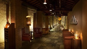 Un pasillo tenuemente iluminado con paredes de piedra y vigas de madera, que incluye sillones de cuero y mesas con velas, evocando un ambiente acogedor y con historia.