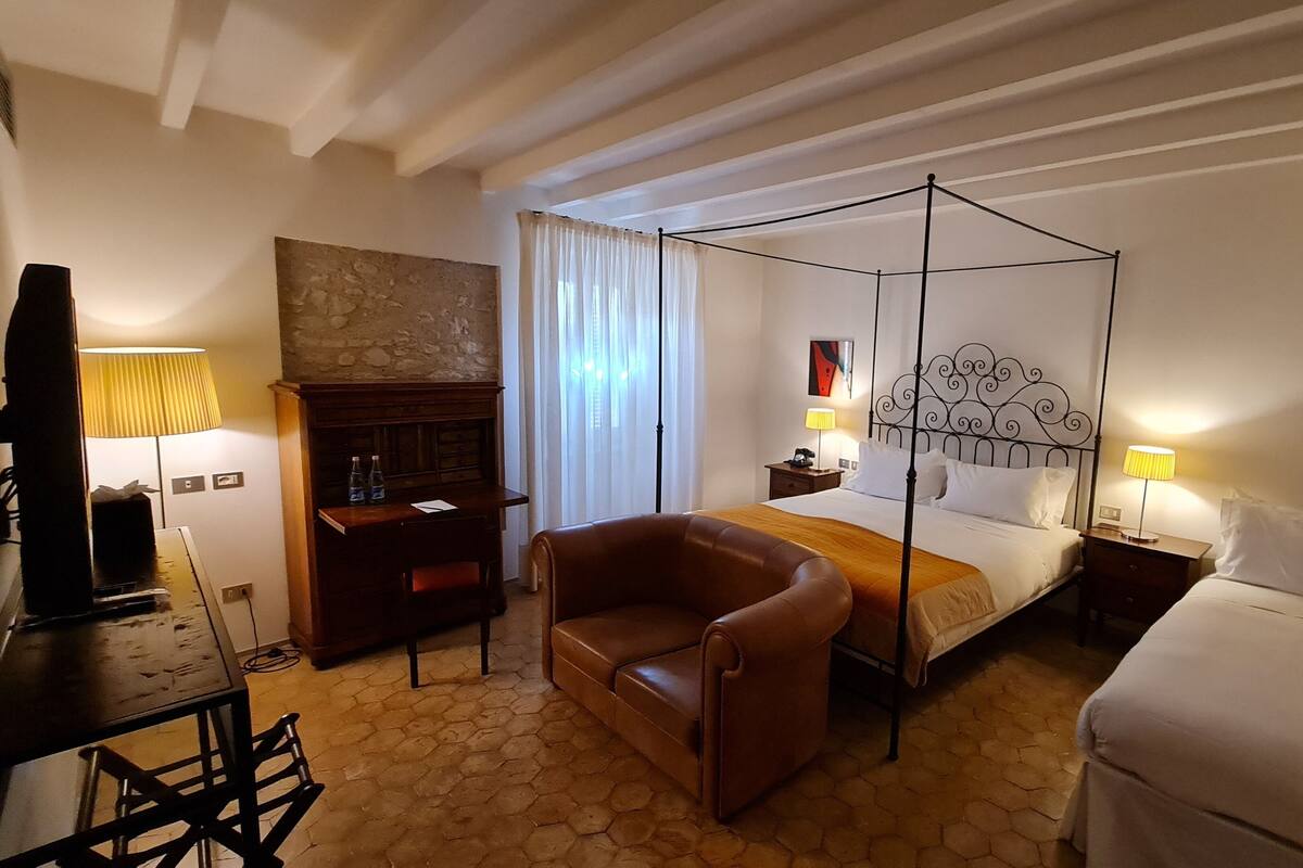 A hotel room with a four-poster bed, a leather armchair, a desk with a lamp, and a minibar.