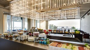 Ein Bereich mit Buffet und vielfältigen Speisen, eine moderne Kronleuchter und große Fenster mit Blick auf die Stadt.
