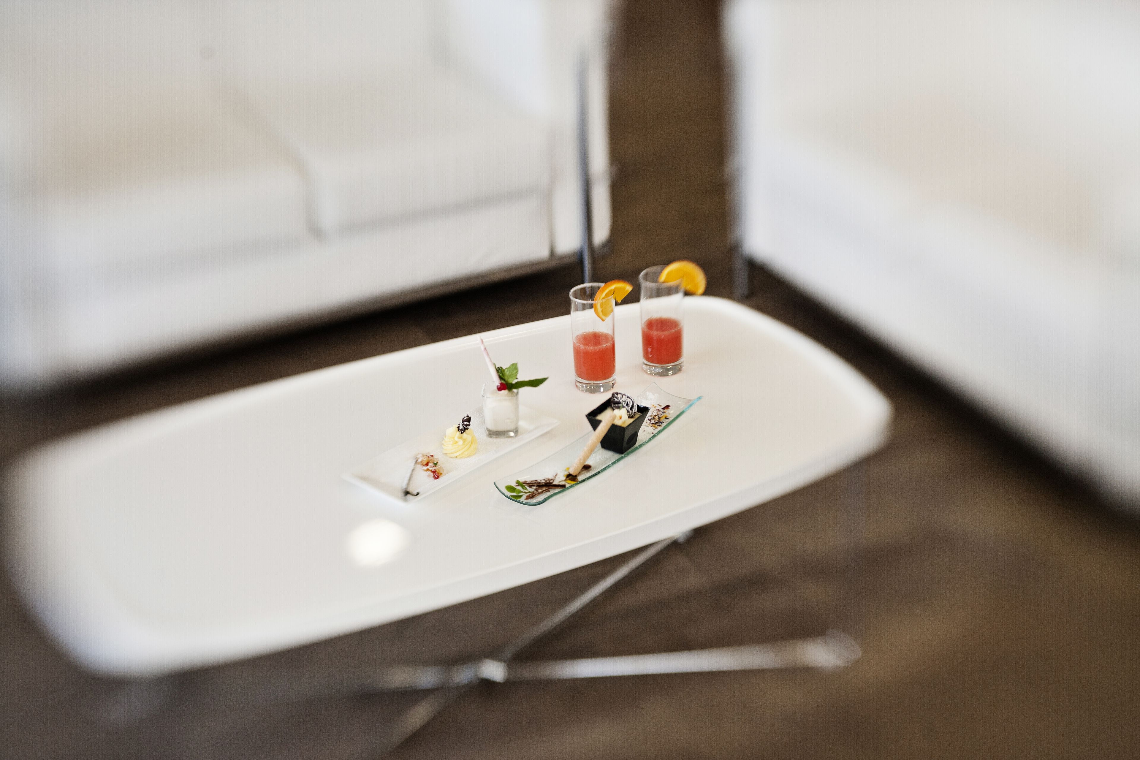 Una bandeja blanca con bebidas y pequeños postres sobre una mesa en un moderno salón con un sofá blanco.