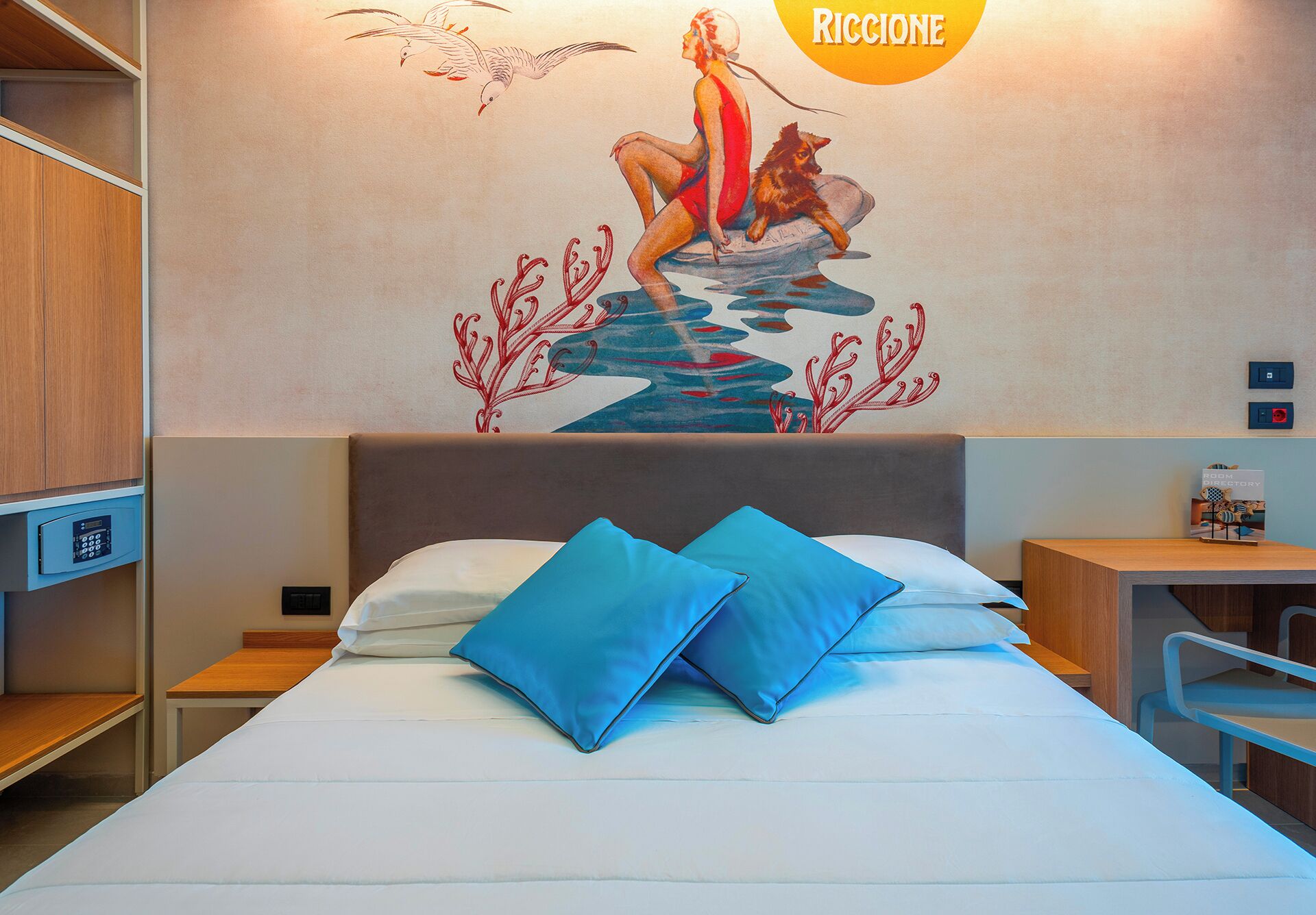 Quarto de hotel com cama, almofadas azuis, um mural na parede, escrivaninha e uma cadeira.