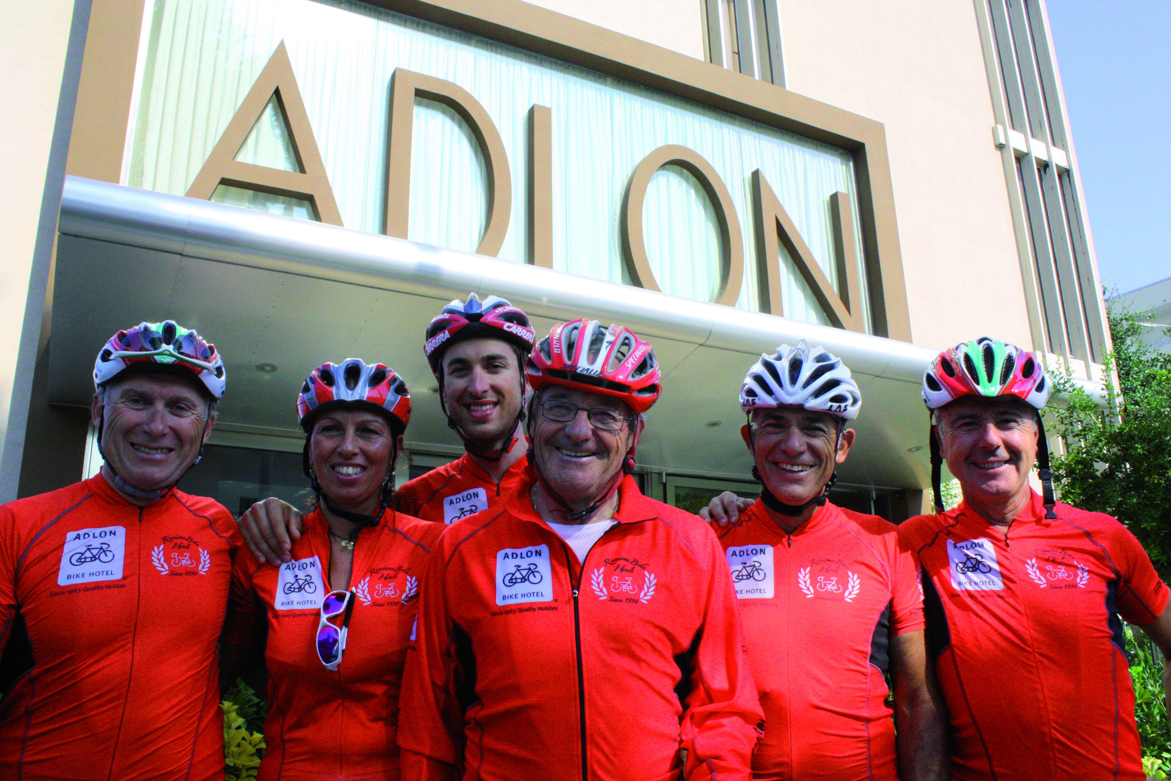 Een groep mensen in rode fietskleding staat voor een gebouw met het bordje 'ADLON'.
