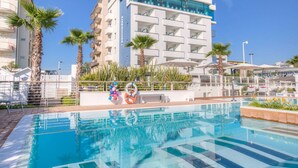 A swimming pool with a clear blue water surface, surrounded by palm trees and a white building with balconies. - Commodore (Riccione)