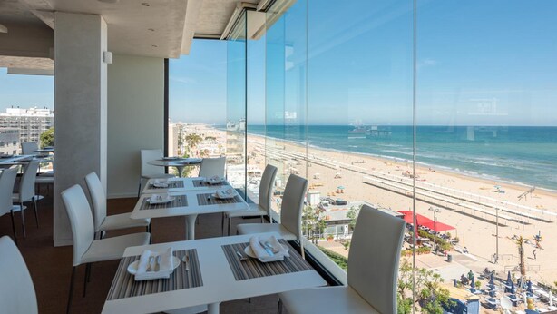 A modern dining area with tables and chairs, large windows offering a view of a beach and ocean. - Commodore (Riccione)