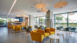 A modern lobby with yellow sofas, grey and yellow chairs, and large windows offering a view of the outdoors. - Commodore (Riccione)