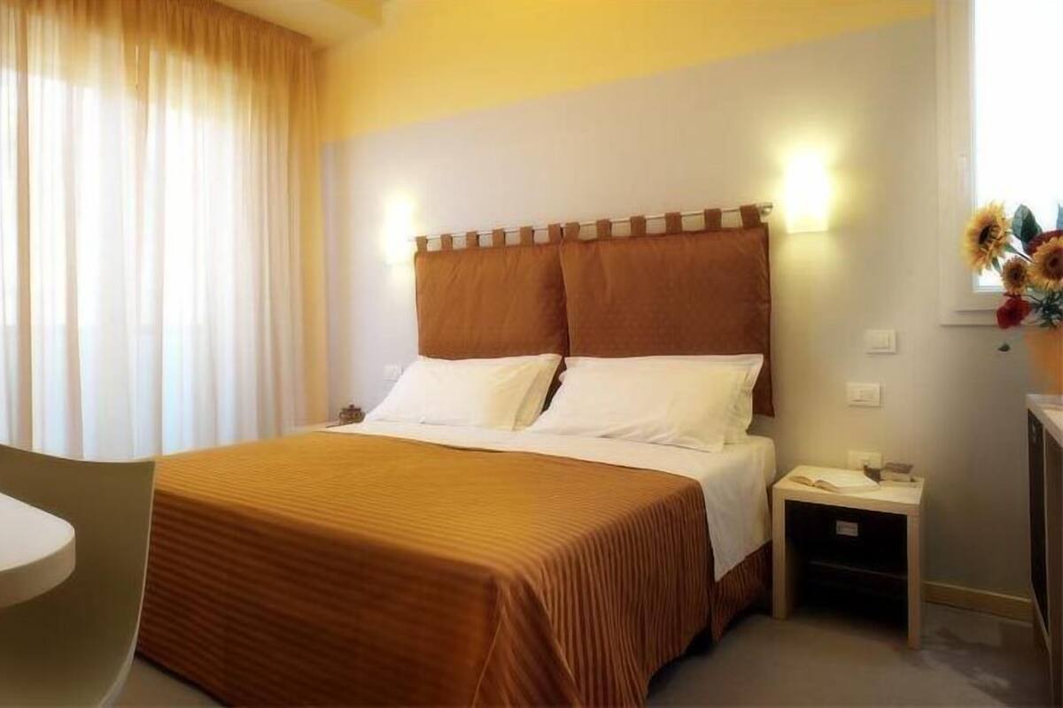 Ein Hotelzimmer mit Bett, Schreibtisch mit Laptop, Nachttisch mit Lampe und einem Fenster mit Vorhängen.
