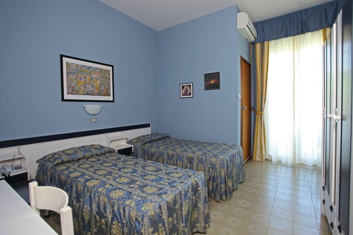 A room with two beds, a desk, and a chair. There is a framed picture on the wall, a window with curtains, and an air conditioning unit.