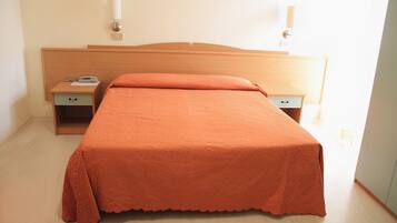 Una camera d'albergo con una testiera in legno, un letto singolo con copriletto arancione, un comodino con il telefono e un piccolo cassetto.