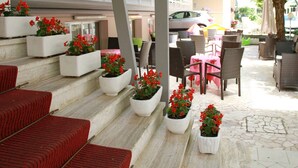 Buiten zitplaatsen met witte plantenbakken vol rode bloemen, rieten stoelen en een roze tafelkleed.