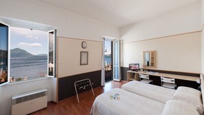 A hotel room with a large window offering a view of the sea and mountains, a desk with a computer, a bed, and a chair.