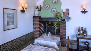 A restaurant with a brick fireplace, green wall, and three framed plates above it. There are tables set with wine glasses and a bench along the wall.