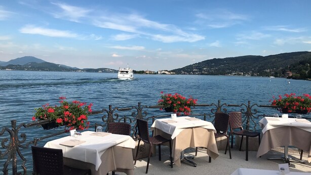 An outdoor dining area with tables and chairs, overlooking a lake with a boat in the distance.