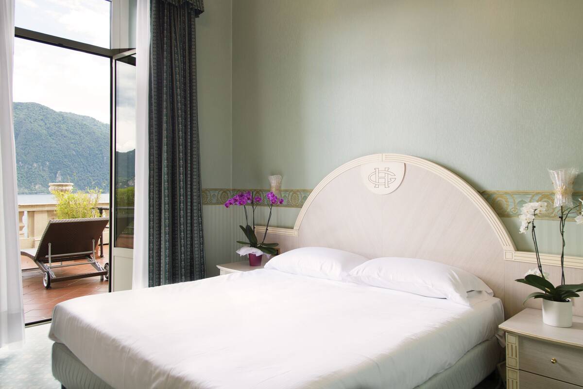 A hotel room with a bed, a nightstand, a window with curtains, a potted plant, and a view of a mountain.