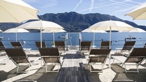 A beachside lounge with sun loungers and umbrellas, overlooking a calm sea and mountains. - Hotel Ghiffa (Ghiffa)