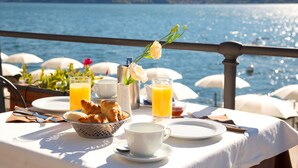 A breakfast table with croissants, a cup, a jug of orange juice, and a vase with a flower, overlooking a lake with umbrellas. - Hotel Ghiffa (Ghiffa)